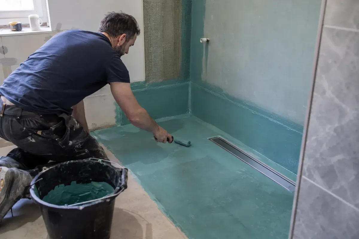 Bathroom installer applying a waterproof tanking membrane with a small roller around a walk-in shower floor and wall junction. Clearly visible linear floor drain, sealed corners and step-free shower zone under construction, waterproofing activity central.