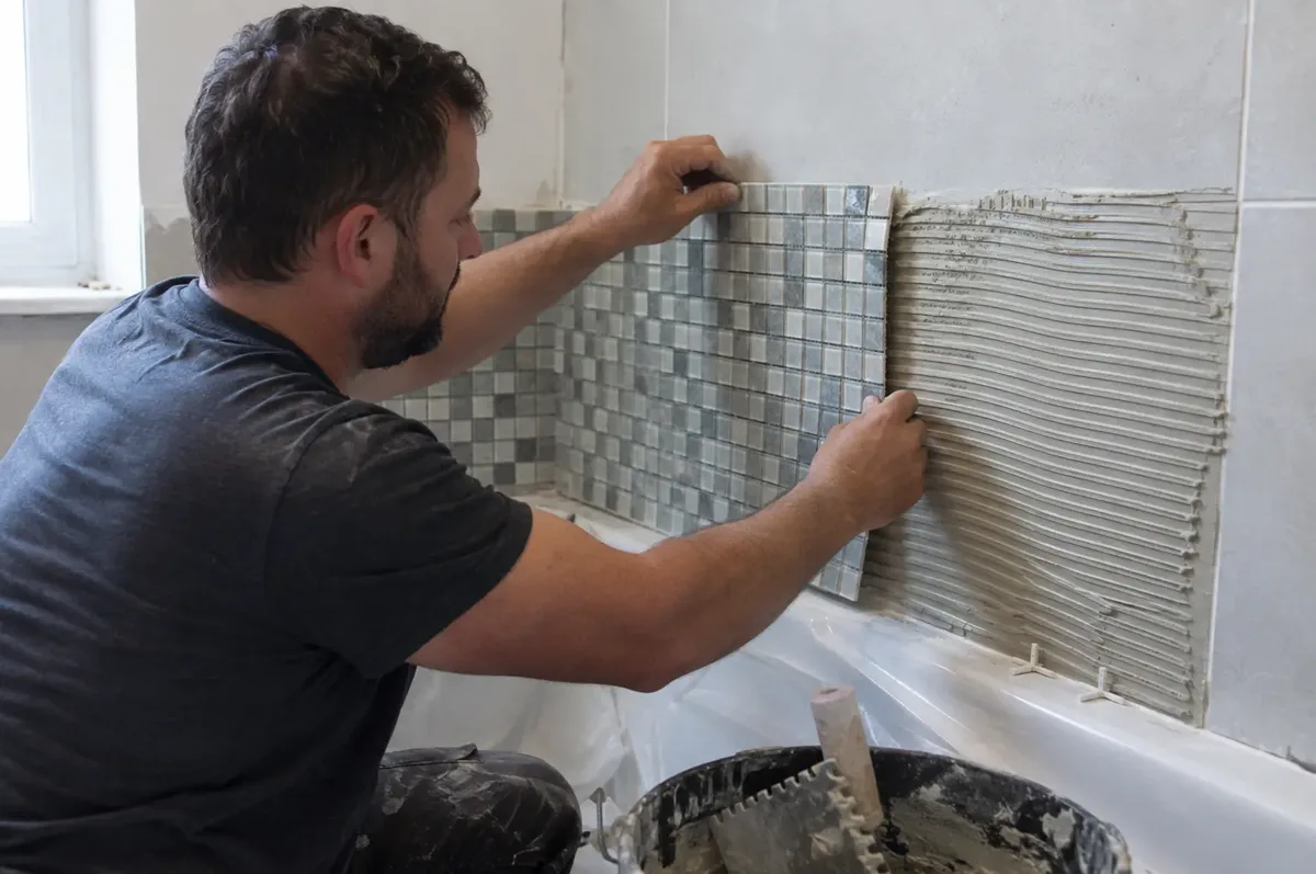 Tiler fixing a sheet of tiny square mosaic tiles to combed adhesive above a bath.