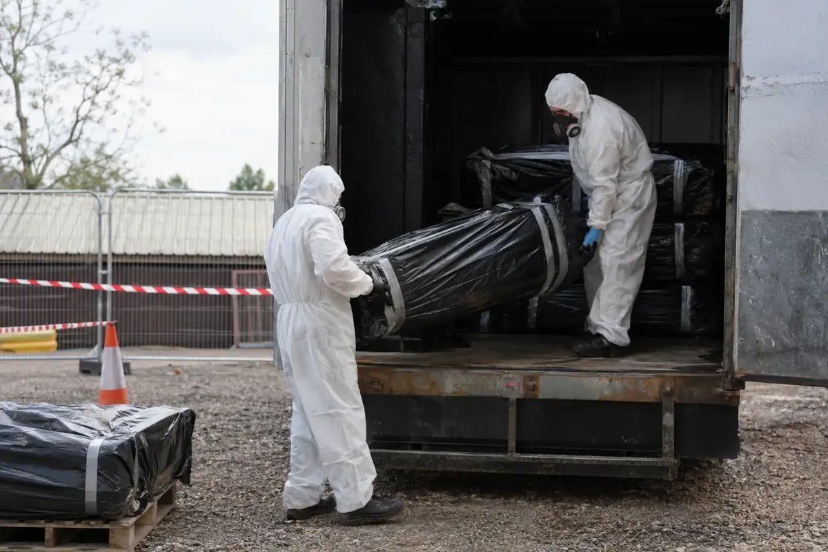 Two asbestos waste specialists in hooded coveralls, respirators and gloves carefully loading fully sealed double-wrapped building-material packages into a dedicated enclosed waste container in a controlled cordoned yard. Packages intact, no loose waste or dust, clear safe handling.